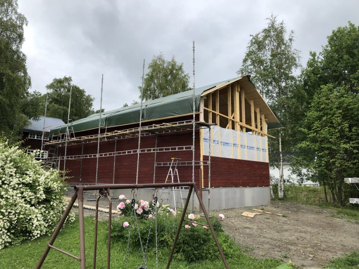 Hus under renovering med ny panel och papp, omgiven av ställning och natur.