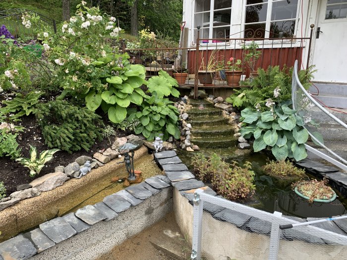 Nybyggd vattenrabbat nära ett hus med plantor, stenkanter och en flyttad reningsdunk i låda.