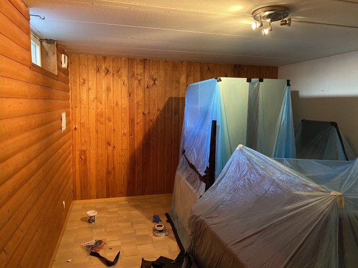 Källarrum under renovering med träpanelväggar och skyddade möbler täckta med plast.