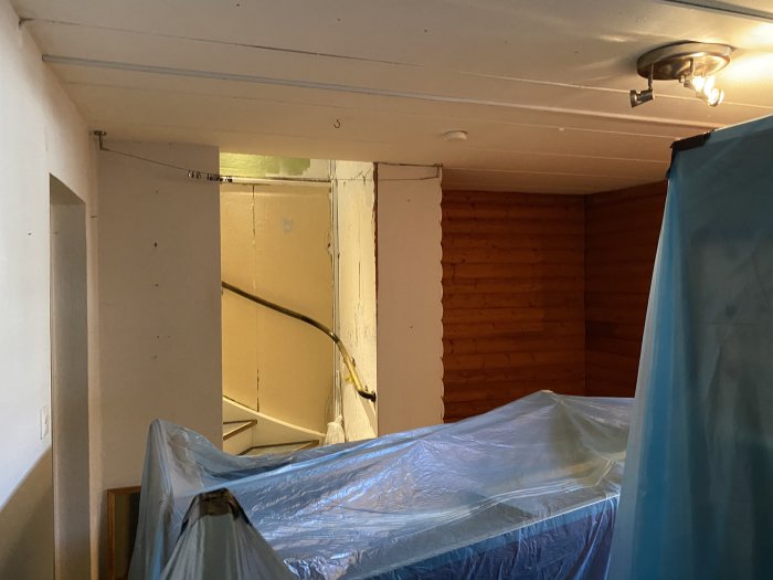 Källarrum under renovering med träpanelväggar och möbler överdragna med blå plastskydd.