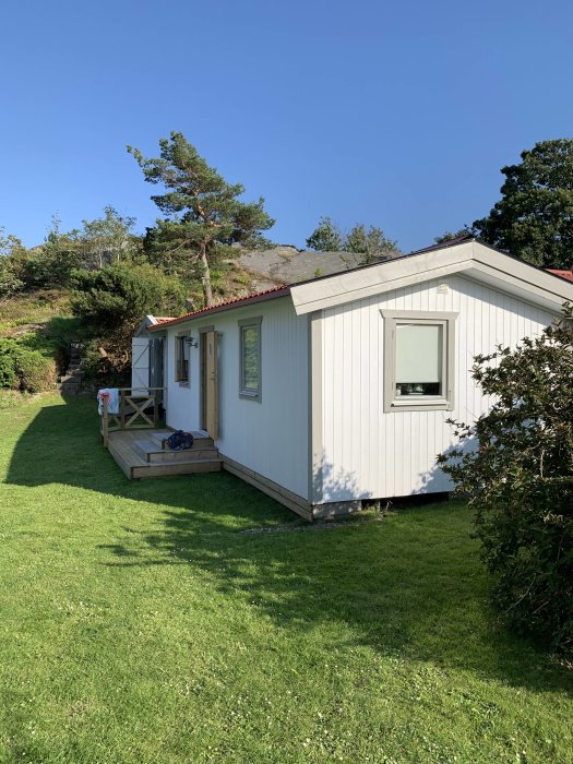 Vit trästuga med veranda på en grönläkt gräsmatta under en klarblå himmel.
