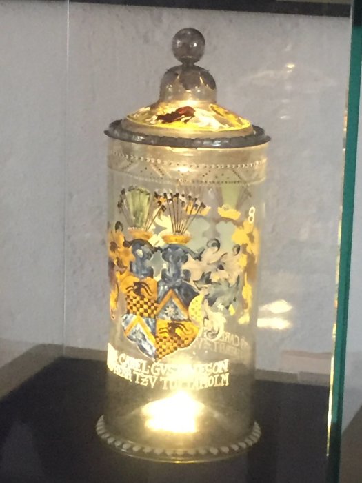 En dekorerad metallbehållare med heraldiska motiv och text, på en hylla bakom glas.