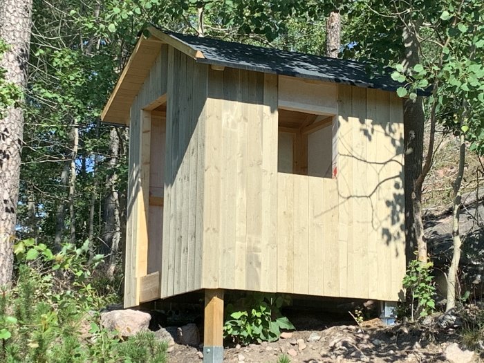 Nybyggd lekstuga i trä som bleknat i solen, omgiven av träd och natur, står på stenar och reglar.