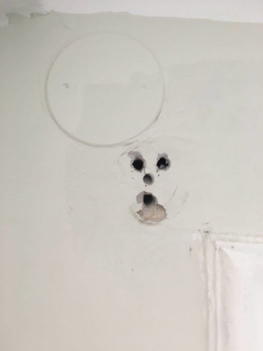 Tre borade hål i en vit vägg med obrukade plastpluggar, ovanför en dörrkarm.