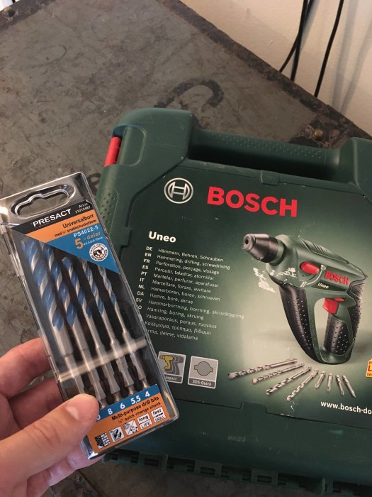 Hand håller förpackning med borr framför öppen Bosch-verktygslåda med borr och skruvdragare.