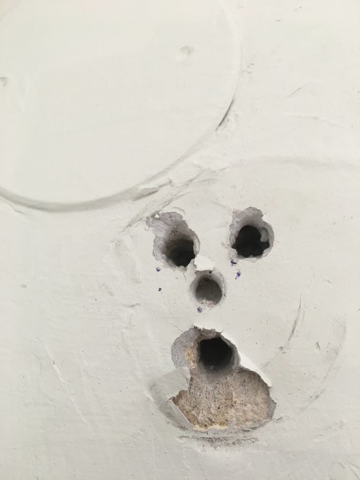 Vit vägg med flera trasiga expansionsbultshål och en oskarp borrhålscirkel.