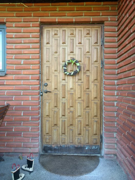 Traditionell träpanel dörr i brun nyans med dekorativ krans, omgiven av röd tegelvägg och två par gummistövlar vid tröskeln.