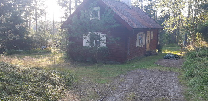 Ett äldre, rött timmerhus med vita knutar på en öppen, kuperad tomt omringad av träd och grönska i dagsljus.