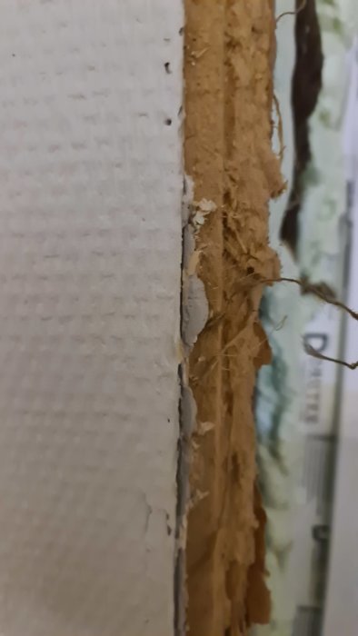 Närbild av en delvis bortriven vävtapet som exponerar spånskiva och träplankor under.