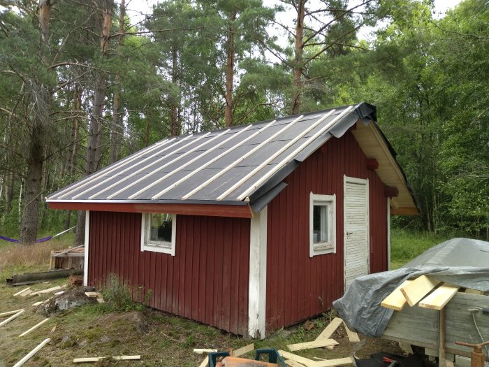 Röd stuga med pågående takläggningsarbete och synlig underlagsduk, omgivet av skog.