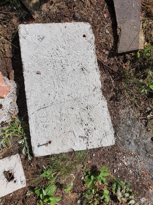 En platta av grå isoleringsmaterial med oregelbundna fibrer ligger på marken med några små löv och en spik på ytan.