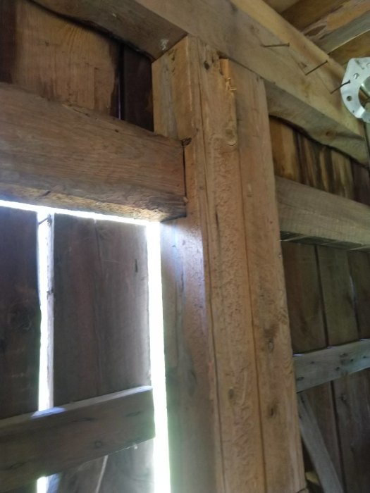 Trästolpe i gammal byggnad med tvärstöttor, ljus sipprar in genom springor i väggpanelen.