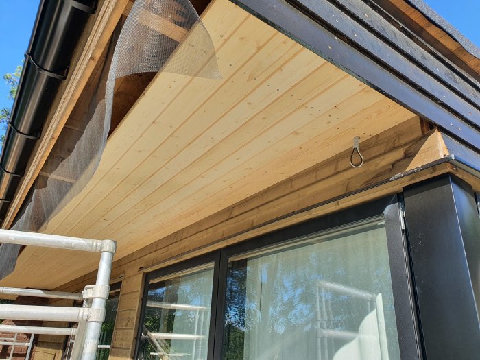 Nyinstallerad ljus träbeklädnad under takfoten på ett hus, med synlig högtalarkabel och L-stöd vid källaringången.