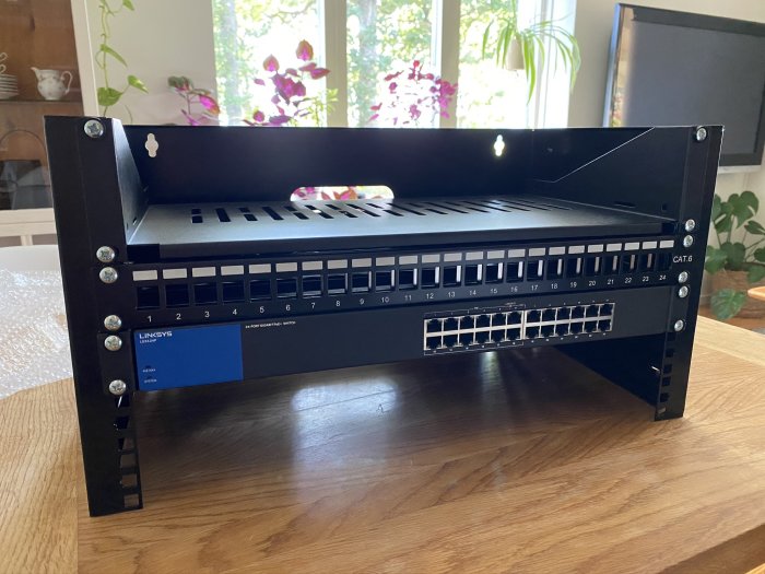 En nätverksswitch och patchpanel monterade i ett rack på ett träbord inomhus, med fönster och blommor i bakgrunden.