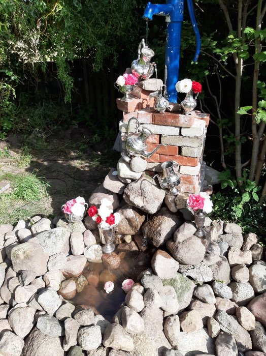 Hemmagjord fontän med stenar, en gammal rullebör som botten, kaffekannor, blommor och en blå pump.