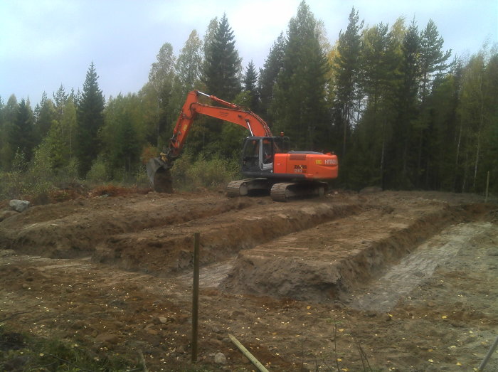 Grävmaskin vid arbete på grunden för ett lösvirkeshus i skogsmiljö.