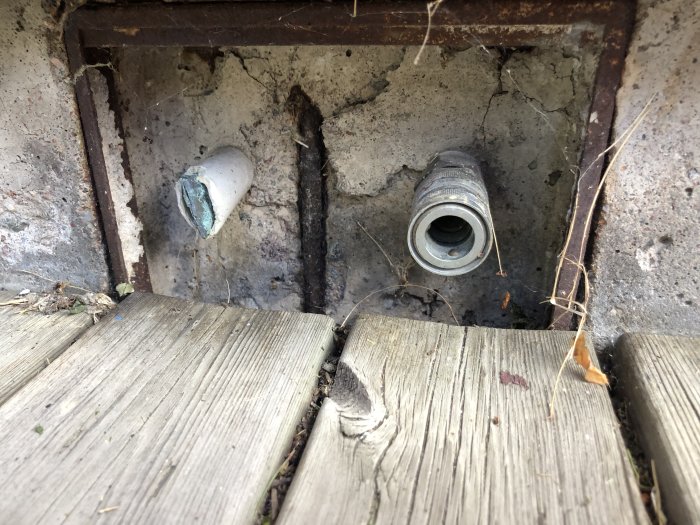 Rostig avloppshål med trasiga rör omgivna av gammalt trägolv och smuts.