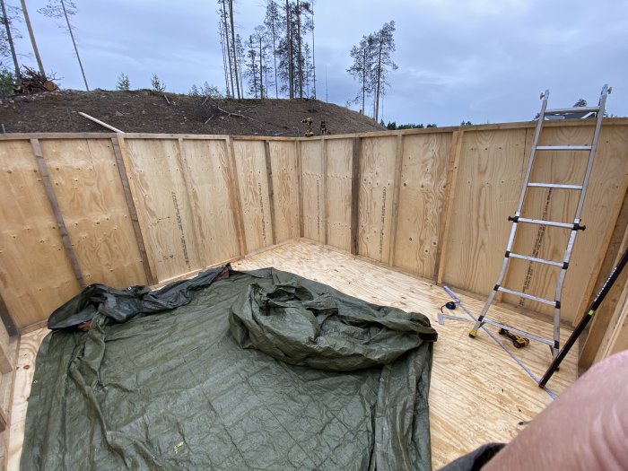 Nybyggt förrådsgolv med uppresade väggsektioner, stege och täckplast, omgivet av skog.