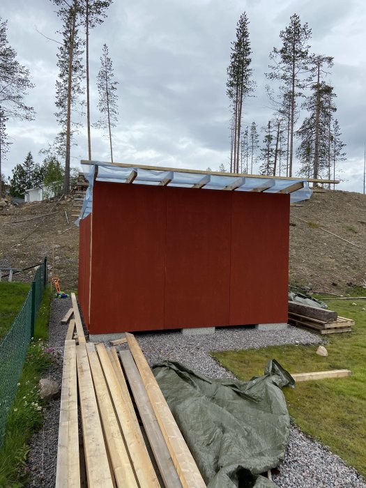 Nybyggt förråd med rödmålade väggar, takstolar och plasttak under konstruktion, med skog i bakgrunden.