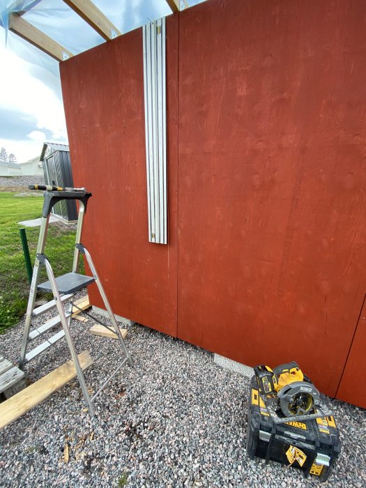 Nymålad förrådsvägg med uppsatta takstolar, stege, och verktyg på grusunderlag.