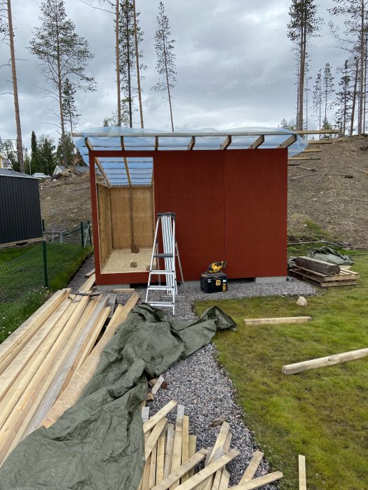 Halvfärdigt förråd med uppresta väggsektioner och takstolar, målat i rött, omgivet av virke och stege.