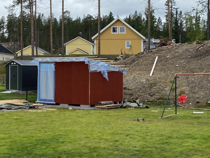 Påbyggt förråd med nymålade röda väggar och blå presenning över oavslutat tak i en trädgård.