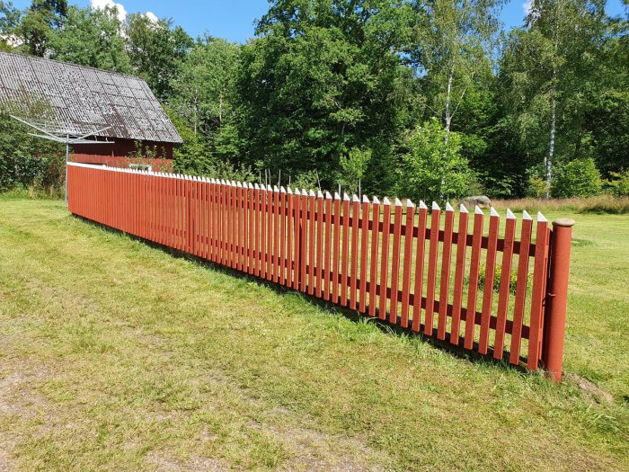 Rött staket av trä framför ett hus med grön gräsmatta och träd i bakgrunden.