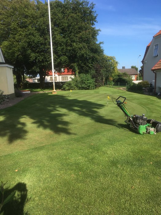 Vertikalskurit gräsmatta med flaggstång och gräsklippare i förgrunden och hus i bakgrunden.