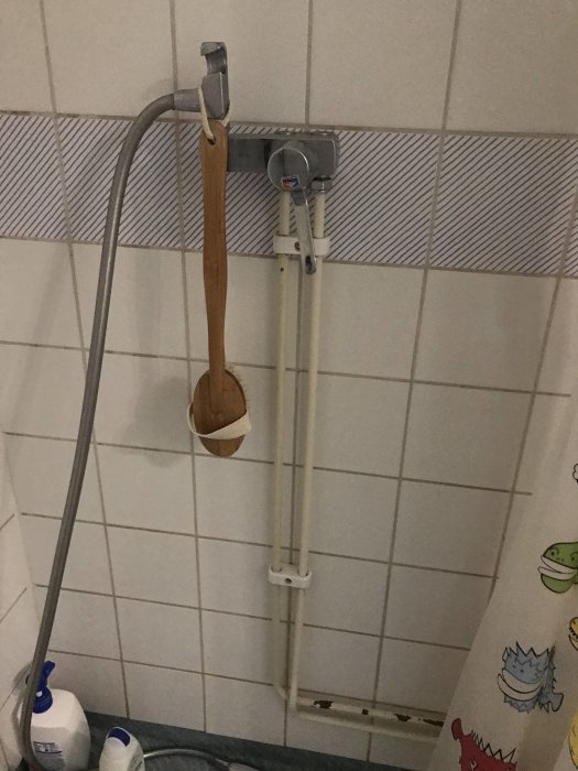 Synliga duschrör längs en kaklad vägg med en duschstång, en träslev och småflaskor i ett badrum.