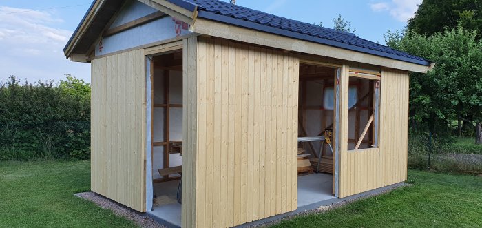 Obyggd friggebod i trä med öppen dörr till vänster och fönster till höger, med trädgården i bakgrunden.