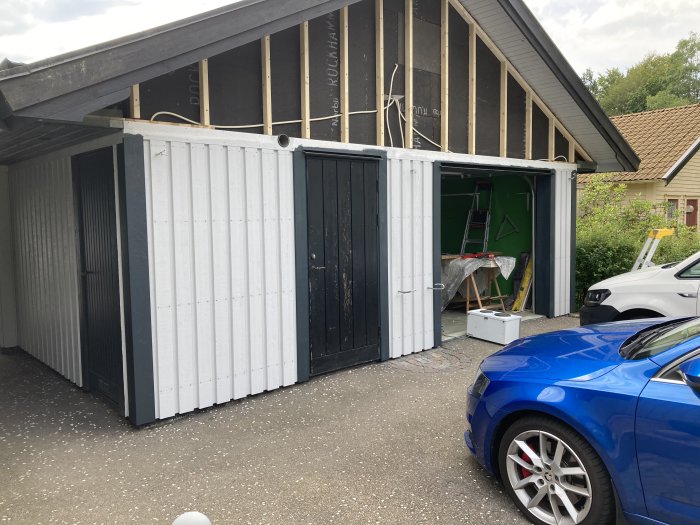 Framsidan av ett hus under renovering med ny stående panel, öppen garageport och material inuti.