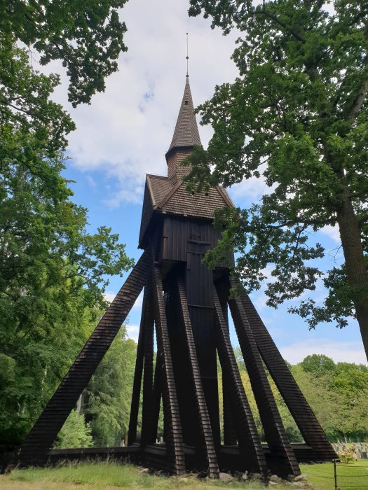 Ett gammalt träkyrkotorn på styltor omgivet av gröna träd, med en blå himmel i bakgrunden.