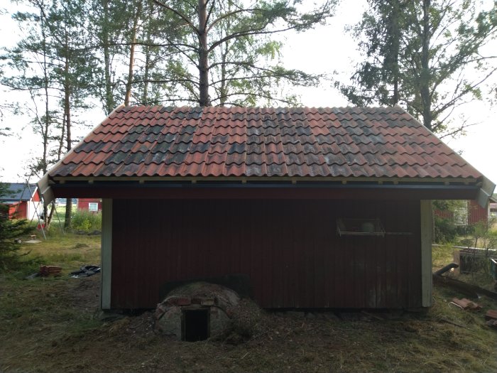 Träfärgad fristående förrådsbyggnad med tegelröd och svart takpannor, omgiven av träd och gräsmark.