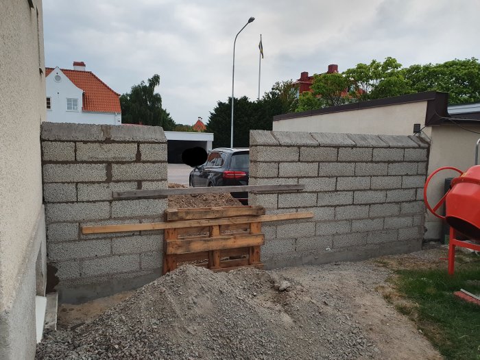 Nybyggd grå mur av betongblock med stödformar mellan ett hus och ett garage, bredvid en grushög.