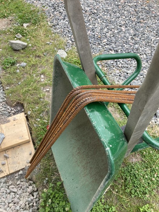 Bockade armeringsjärn i olika storlekar vilande på en grön skottkärra mot en grusig bakgrund.