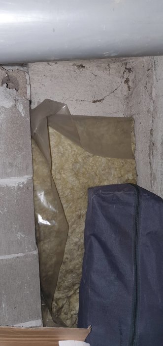 Oisolerat hörn av ett litet rum med synlig glasull och plast, sprickor i betong och en del av en mörk väska.