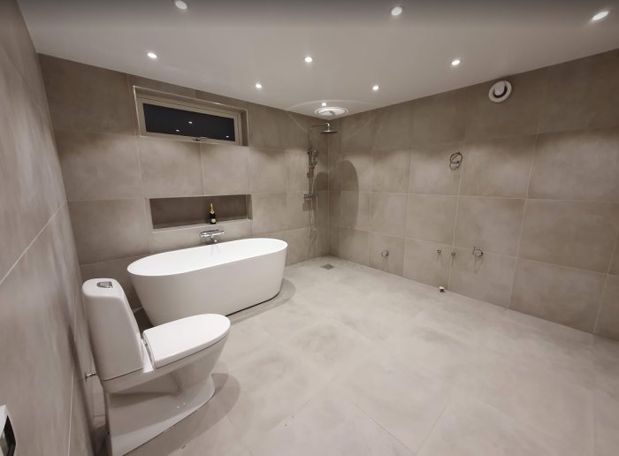 Modernt badrum med frittstående badkar, dusch, toalett och vägg i kakel.