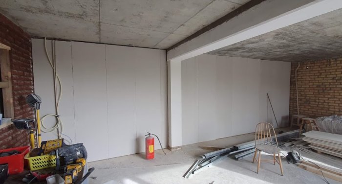 Renoveringsarbete med nya fibercementskivor på väggen i ett rått rum med byggmaterial och verktyg.