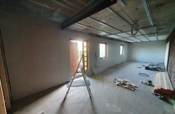 Renoveringsarbete i ett tomt rum med OSB-paneler på väggarna och synliga takreglar, stege och byggmaterial är synligt.