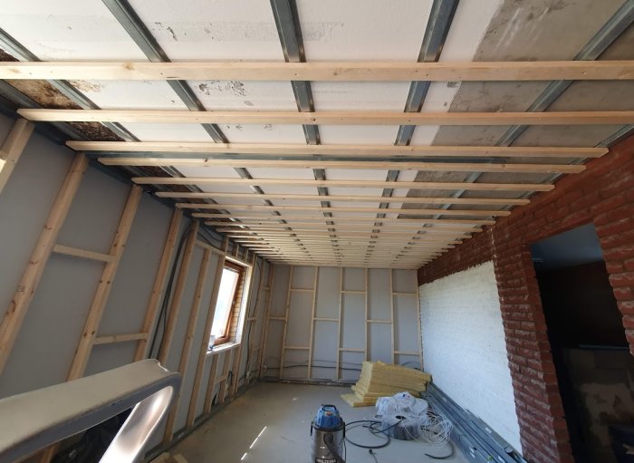Renoveringsarbete med exponerade trä- och plåtprofiler i ett tak och tegelväggar i ett tomt rum under ombyggnad.
