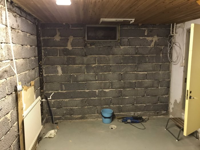Orenoverad källartvättstuga med gråmålat golv, gråa betongblocksväggar, delvis synlig radiator och synliga rör och ledningar.
