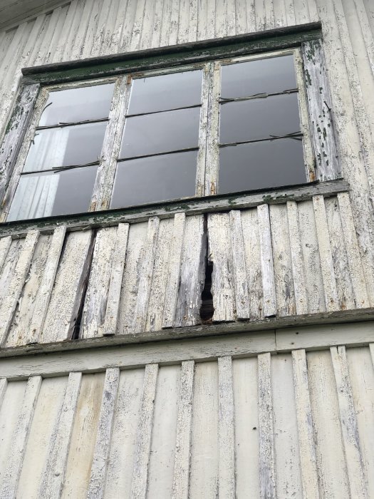 Slitet hus med fuktskador, visar vitmålad fasad, fönster med sprucket glas och flagande färg.