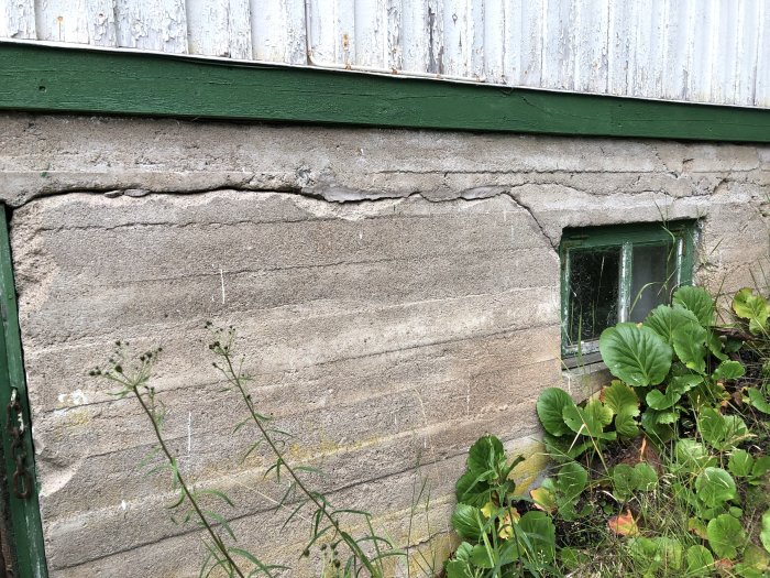 Fuktskador på en betongvägg med sprickor och avflagning nedanför ett slitet metalltak och vid ett litet fönster.