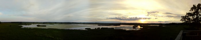 Panorama av landskap vid solnedgång med öppet fält, vatten och träd mot en färgrik himmel.