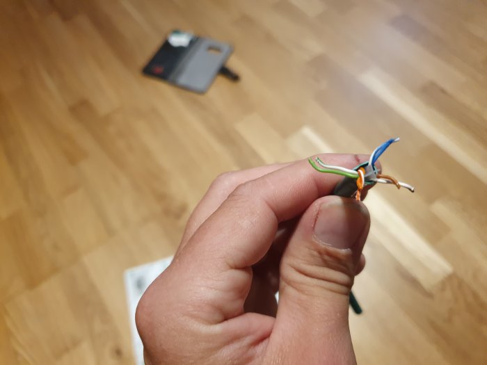 Hand som håller nätverkskabel med korsande gröna och orangea par mot ett trägolv-bakgrund.