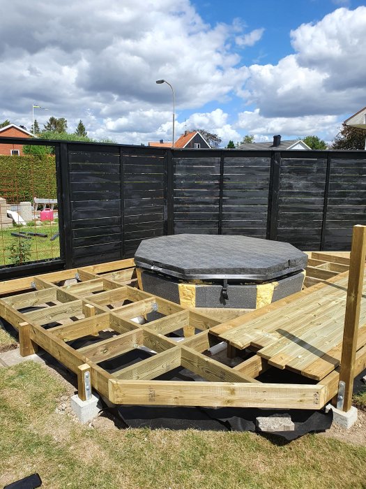 Pågående bygge av träaltan runt en bubbelpool, med svart staket i bakgrunden under en molnig himmel.