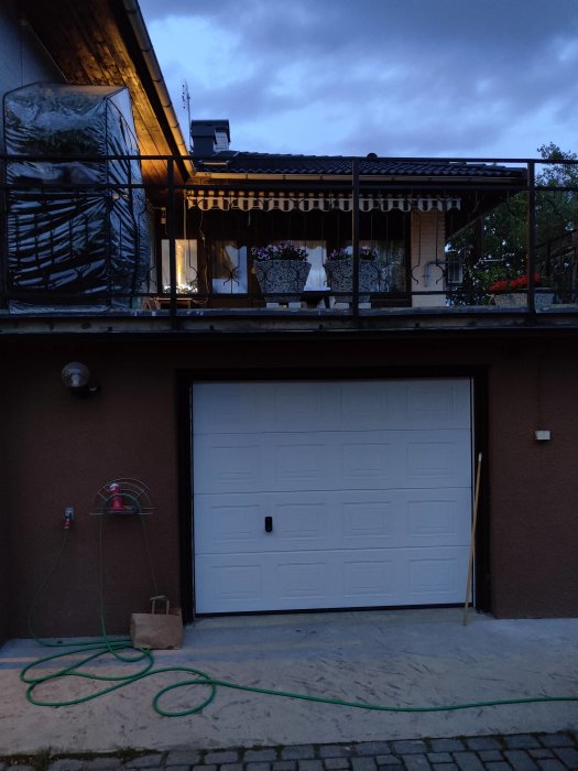 Kvällsbild av en byggnad med garageport i förgrunden och en terrass ovanpå med klinkers och blomkrukor.