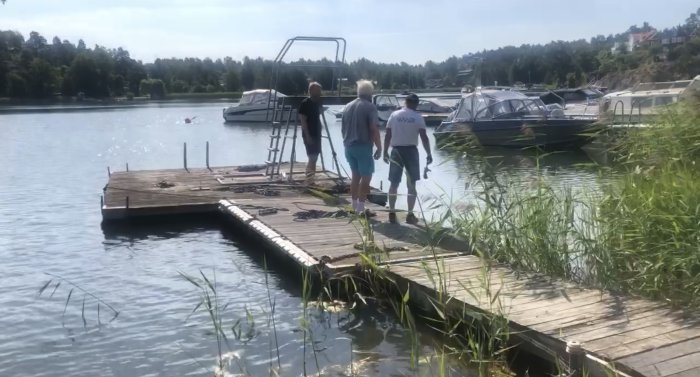 Personer på en renoverad flotte vid en badplats, med hopptorn och båtar i bakgrunden.