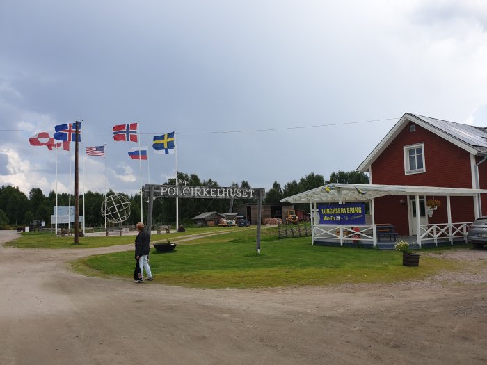 En person går förbi en röd byggnad med skylten "Lunchservering" och flera nationella flaggor framför skulpturen av en jordglob vid Polcirkeln.
