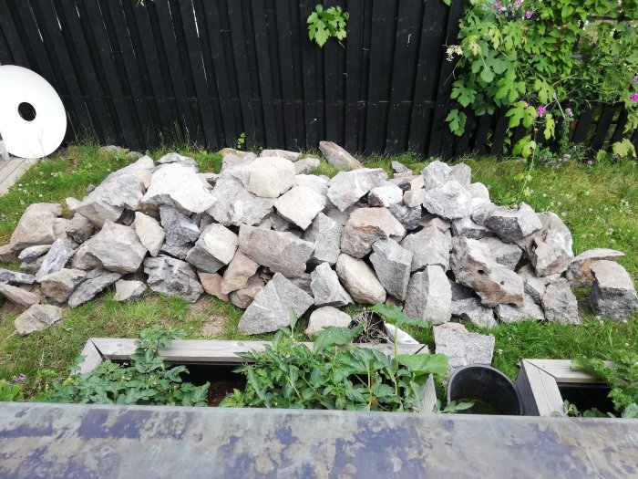 Hög med blandade stenblock på gräsmatta framför ett svart staket och gröna växter.
